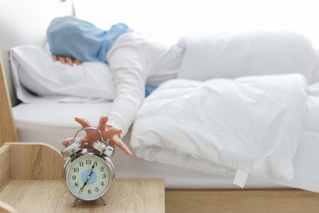 Oversleeping asian woman wearing white muslim sleepwear lying on bed, missing ring of alarm clock waking up. Rear view portrait young girl hand trying to stop ringing while her face down on pillowの写真素材