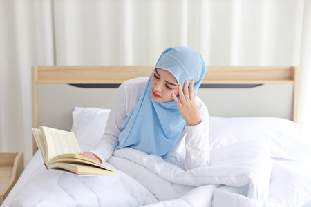 Happy young beautiful asian woman in muslim sleepwear sitting, reading book on bed. Cheerful and cute girl resting in bedroom and copy space for your advertising content.の写真素材
