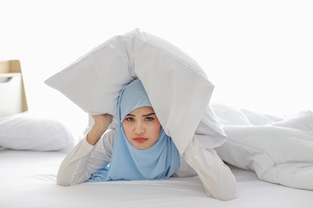 Oversleeping asian woman in muslim sleepwear lying on bed and missing ring of alarm clock waking up. Young girl trying to sleep by putting pillow over her head in bedroom with unhappy emotionの写真素材