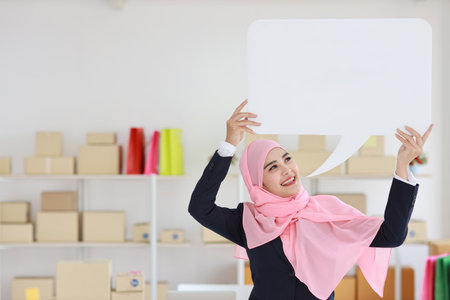 Active asian woman in muslim blue suit standing and holding white speech bubble with online package box delivery background. Startup small business SME freelance girl work at home with happy face.の写真素材