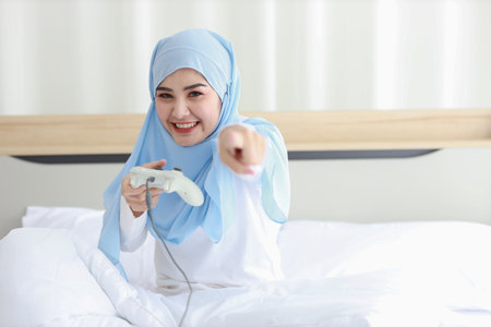 VDO game console station concept. active asian woman wearing white muslim sleepwear sitting on bed, holding joystick and playing exciting game. Cute girl looked excited with game controller consoleの写真素材