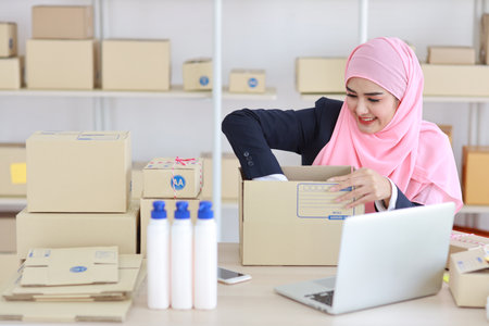 Active smiling asian woman in muslim suit sitting and working with online package box delivery. Startup small business SME freelance girl working on computer and mobile phone with happy face.の写真素材