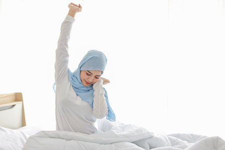 Beautiful asian woman stretching her arms after getting up in the morning on bed at sunrise in bedroom. Cute young girl in muslim dress and hijab sitting and relaxing with happy and smiling face.の写真素材