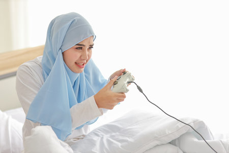 VDO game console station concept. active asian woman wearing white muslim sleepwear sitting on bed, holding joystick and playing exciting game. Cute girl looked excited with game controller consoleの写真素材
