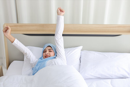 Beautiful asian woman wearing white muslim sleepwear lying on bed, stretching her arms after getting up in the morning at sunrise. Cute young girl with hijab wake up and relaxing, closing her eyesの写真素材