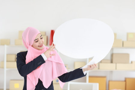 Active asian woman in muslim blue suit standing and holding white speech bubble with online package box delivery background. Startup small business SME freelance girl work at home with happy face.の写真素材