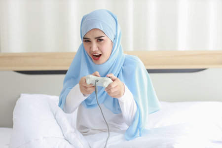 VDO game console station concept. active asian woman wearing white muslim sleepwear sitting on bed, holding joystick and playing exciting game. Cute girl looked excited with game controller consoleの写真素材