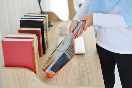 Closeup active young asian muslim housewife woman hands cleaning with vacuum cleaning wooden table with computer blurred background. Cleaning girl in white dress and black leggings vacuuming her roomの写真素材