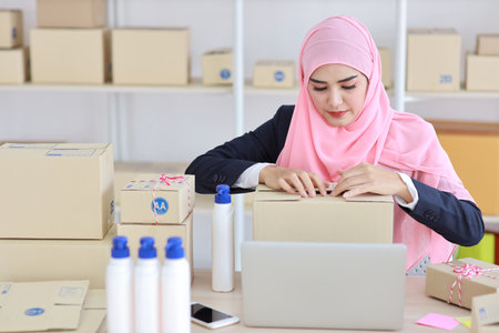 Active smiling asian woman in muslim suit sitting and working with online package box delivery. Startup small business SME freelance girl working on computer and mobile phone with happy face.の写真素材