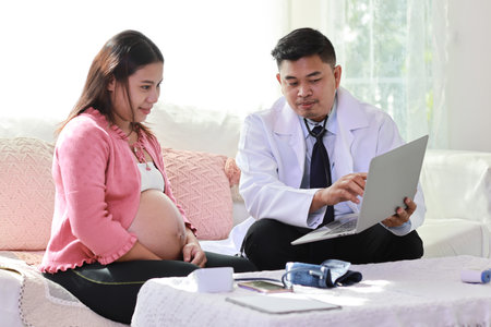 Happy smiling young asian pregnant female and male doctor sitting on sofa in living room at home with private treatment and consultant. Friendly doctor showing result from computerの写真素材