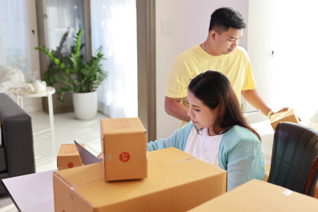 Asian couple working on box packaging, attractive young asian pregnant woman sitting and using tablet, mobile smart phone while writing order from customer with computer and husband working togetherの写真素材