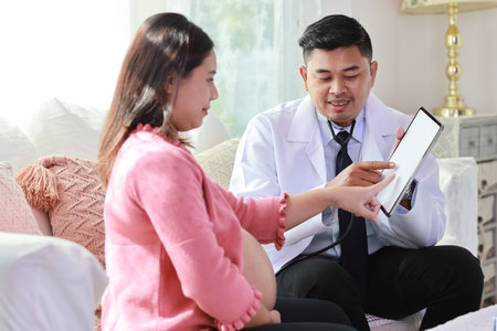 Happy smiling young asian pregnant female and male doctor sitting on sofa in living room at home with private treatment and consultant. Friendly doctor showing result from tabletの写真素材