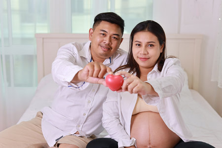 Happy asian adult couple with husband sitting and resting on bed in bedroom while holding heart symbol and smile. Expectant mother preparing and waiting for baby birth during pregnancy.の写真素材