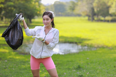 Young beautiful asian volunteer woman hold garbage bag and picking up litter while cleaning plastic bottle from lawn and showing full black bag during a volunteer environmental cleanup area park eventの写真素材