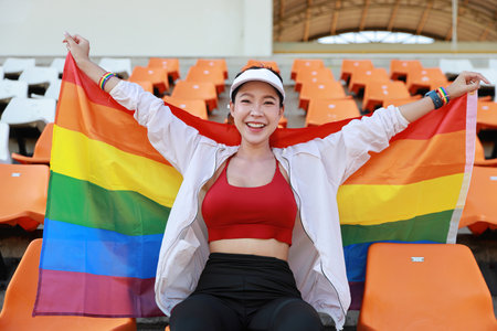 Happy and excited young female asian transgender LGBT sport fan cheering and watching the match with rainbow flag raising and wristband, she sitting amongst the rows of empty seats on stadiumの写真素材