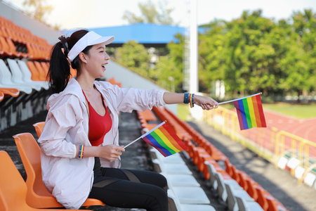 Happy and excited young female asian transgender LGBT sport fan cheering and watching the match with rainbow flag pointing and wristband, she sitting amongst the rows of empty seats on stadiumの写真素材
