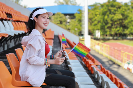 Happy and excited young female asian transgender LGBT sport fan cheering and watching the match with rainbow flag raising and wristband, she sitting amongst the rows of empty seats on stadiumの写真素材