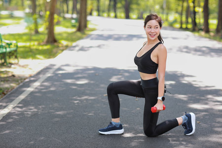 Strong and beautiful asian fitness female sportswoman lifting dumbbell weight training after finish morning run outdoor. Happy smiling athlete woman doing squats exercise under trees in the parkの写真素材