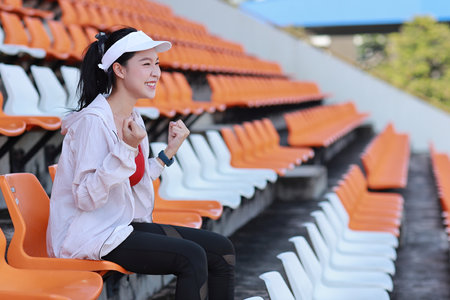Portrait of happy and excited young female asian sport fan cheering and watching the match with her hands clapping, she sitting amongst the rows of empty seats on stadium while celebrate winner teamの写真素材