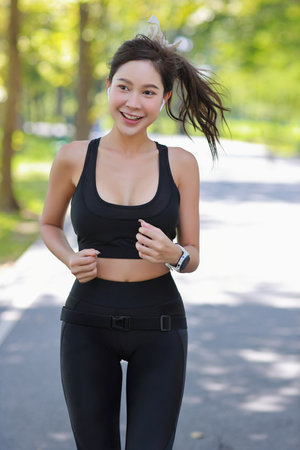Beautiful asian fitness female sportswoman runner doing morning jogging exercise training outdoor. Happy smile athlete woman running under the tree in the park while looking at camera and listen musicの写真素材
