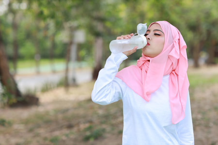 Beautiful fitness athlete asian woman drinking water after work out exercising. Young cute girl standing in muslim sportswear with headphone taking rest after workout outdoor.  Healthy, sport conceptの写真素材