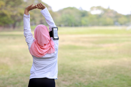 Public exercise lifestyle concept, fitness woman listening music from wireless earphones and mobile phone. Rear view athletic young asian girl in muslim sportswear standing, stretching after workoutの写真素材