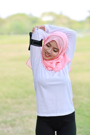 Public exercise lifestyle concept, fitness woman listening music from wireless earphones and mobile phone.  Athletic young asian girl in muslim sportswear standing, stretching after workoutの写真素材
