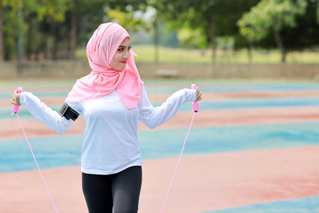 Athletic young asian woman in muslim sportswear standing and skipping rope outdoor for morning exercise. Active girl work out on biceps exercise with green tree background. Sport conceptの写真素材