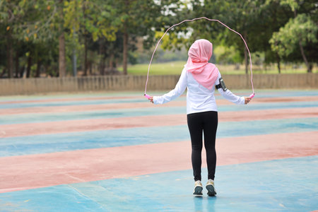 Rear view athletic young asian woman in muslim sportswear standing and  skipping rope outdoor for morning exercise. Active girl work out on biceps exercise with green tree background. Sport conceptの写真素材