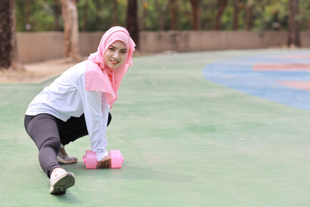Athletic young beauty asian woman in muslim sportswear stretching and lifting dumbbells public exercise. Active girl work out on biceps exercise with green tree background. Sport conceptの写真素材