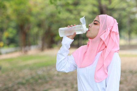 Beautiful fitness athlete asian woman drinking water after work out exercising. Young cute girl standing in muslim sportswear with headphone taking rest after workout outdoor.  Healthy, sport conceptの写真素材