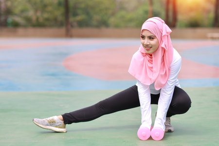 Athletic young beauty asian woman in muslim sportswear stretching and lifting dumbbells public exercise. Active girl work out on biceps exercise with green tree background. Sport conceptの写真素材