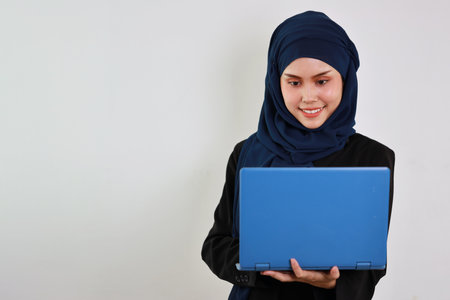 Happy smart asian business woman in muslim dress standing while using computer and smiling with confident in studio. Isolated white background portrait with beautiful face in hijab.の写真素材