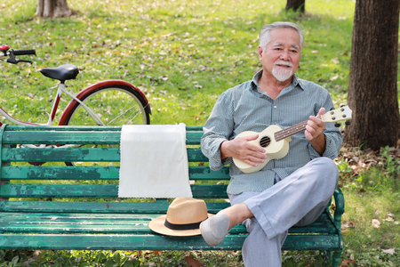 Happy smiling asian senior man with beard sitting on bench playing ukulele and singing a song in garden park outdoor. Musical and relaxation makes elder male happiness. Health care lifestyle concept.の写真素材