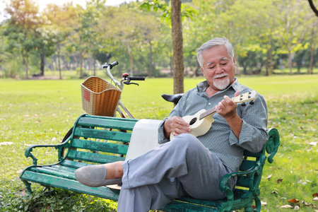 Happy smiling asian senior man with beard sitting on bench playing ukulele and singing a song in garden park outdoor. Musical and relaxation makes elder male happiness. Health care lifestyle concept.の写真素材
