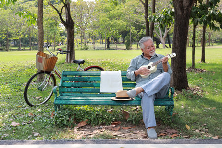 Happy smiling asian senior man with beard sitting on bench playing ukulele and singing a song in garden park outdoor. Musical and relaxation makes elder male happiness. Health care lifestyle concept.の写真素材