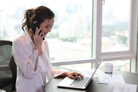 Portrait of confident and beautiful young asian businesswoman with happy face sitting and using computer and mobile smartphone in hotel with city background. Success business and lifestyle conceptの写真素材