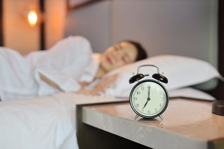 Alarm clock with blurred image of oversleeping woman wearing white sleepwear lying or sleeping on bed in bedroom with happiness and smile. Lifestyle conceptの写真素材