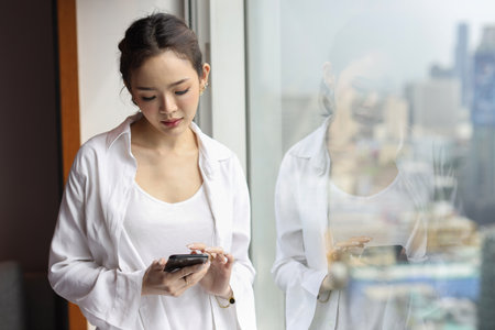 Portrait of confident and beautiful young asian businesswoman with happy emotion standing and using smartphone in modern hotel with city blurred background. Success business and lifestyle conceptの写真素材