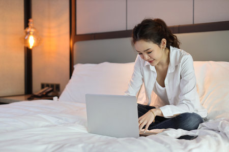 Active asian woman in casual sitting on bed and working on computer with notebook in hotel, beauty businesswoman with bedroom background. Lifestyle technology conceptの写真素材