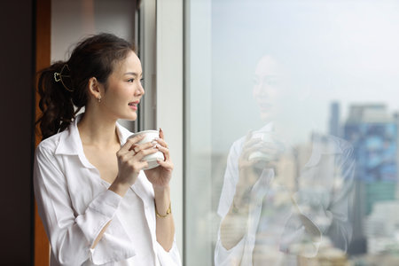 Portrait of confident and beauty young asian businesswoman with coffee cup standing and looking away in modern hotel with city blurred background. Success business and lifestyle conceptの写真素材