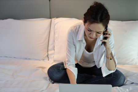 Active and unhappy asian woman in casual sitting on bed and working smart mobile phone and computer in hotel, beauty businesswoman with bedroom background. Lifestyle technology conceptの写真素材