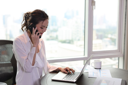 Portrait of confident and beautiful young asian businesswoman with happy face sitting and using computer and mobile smartphone in hotel with city background. Success business and lifestyle conceptの写真素材