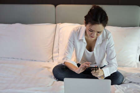 Active and happy asian woman in casual sitting on bed and working smart mobile phone and computer in hotel, beauty businesswoman with bedroom background. Lifestyle technology conceptの写真素材