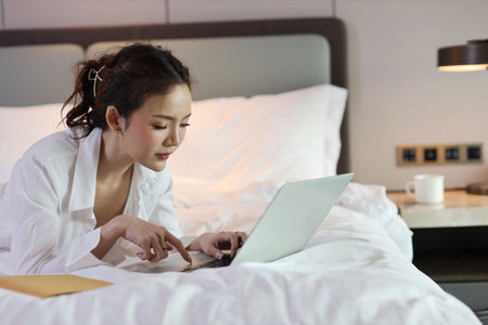 Active asian woman in casual lying on bed and working on computer with notebook in hotel, beauty businesswoman with bedroom background. Lifestyle technology conceptの写真素材