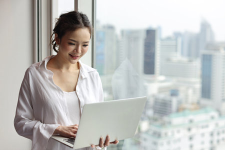 Portrait of confident and beautiful young asian businesswoman with happy emotion standing and using computer in modern hotel with city blurred background. Success business and lifestyle conceptの写真素材