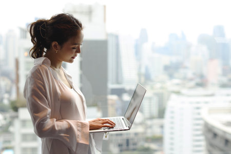 Portrait of confident and beautiful young asian businesswoman with happy emotion standing and using computer in modern hotel with city blurred background. Success business and lifestyle conceptの写真素材
