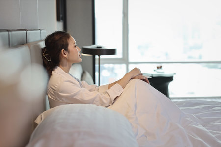 Young depressed asian woman in casual dress sitting and looking at the ceiling while crying on bed in hotel bedroom with low light environment. Sad girl, unhappy and loneliness concept.の写真素材