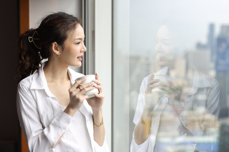 Portrait of confident and beauty young asian businesswoman with coffee cup standing and looking away in modern hotel with city blurred background. Success business and lifestyle conceptの写真素材