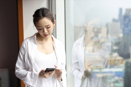 Portrait of confident and beautiful young asian businesswoman with happy emotion standing and using smartphone in modern hotel with city blurred background. Success business and lifestyle conceptの写真素材